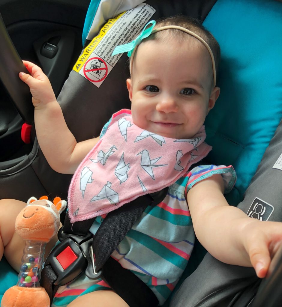 A cleft palate baby riding in her car seat, wearing a pink origami-patterned bandanna bib.