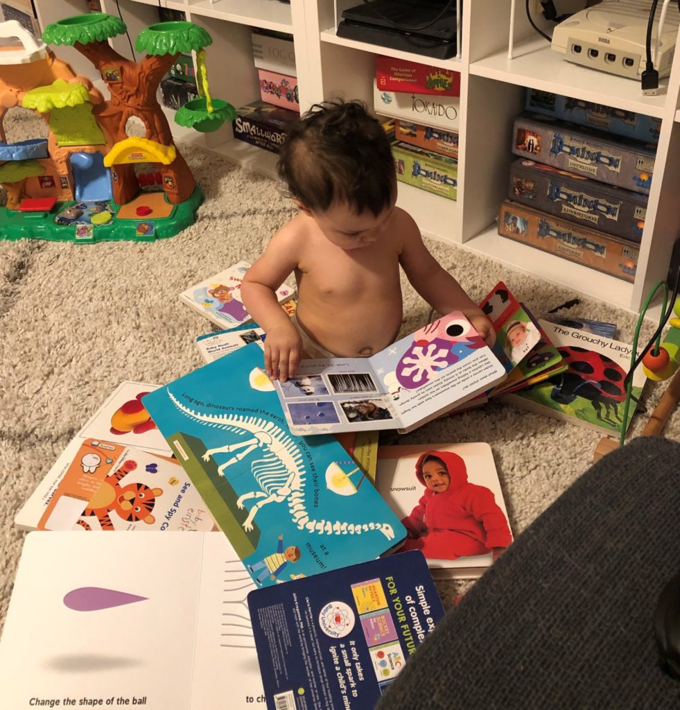 A shirtless toddler surrounded by books, reading a book in her lap