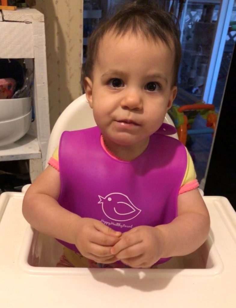 A toddler girl wearing a purple bib and doing sign language for the word "more"