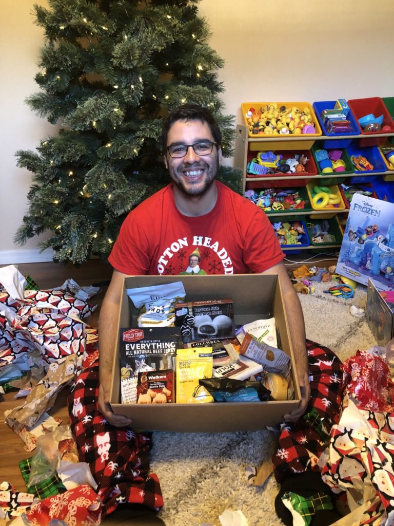 A man sitting in front of a Christmas tree smiling, holding a box full of snack items