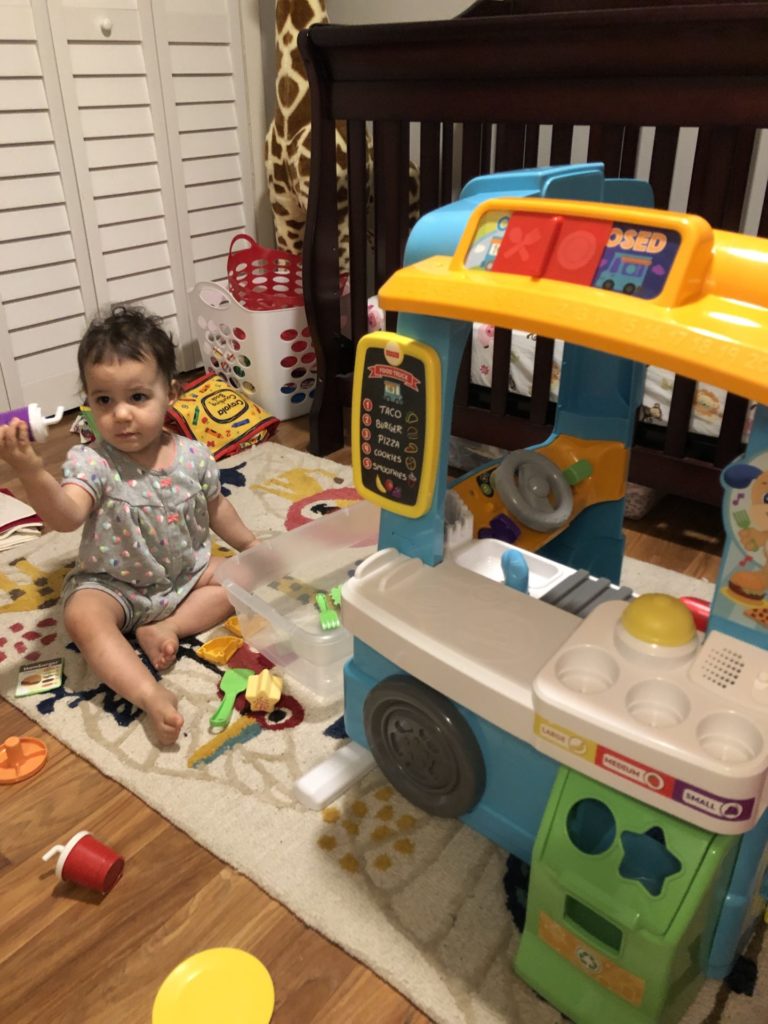 A toddler girl wearing a grey romper playing with a toy food truck
