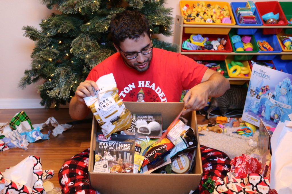 A man sitting in front of a Christmas tree surrounded by wrapping paper, looking through a box full of snack items
