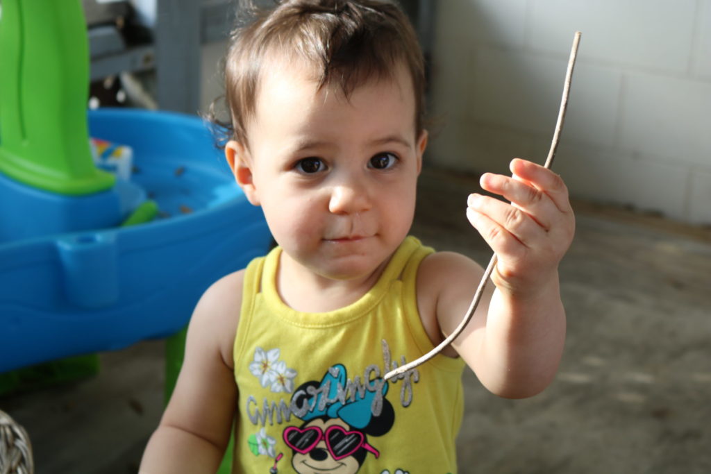 A toddler in a yellow shirt holding a stick up for the camera