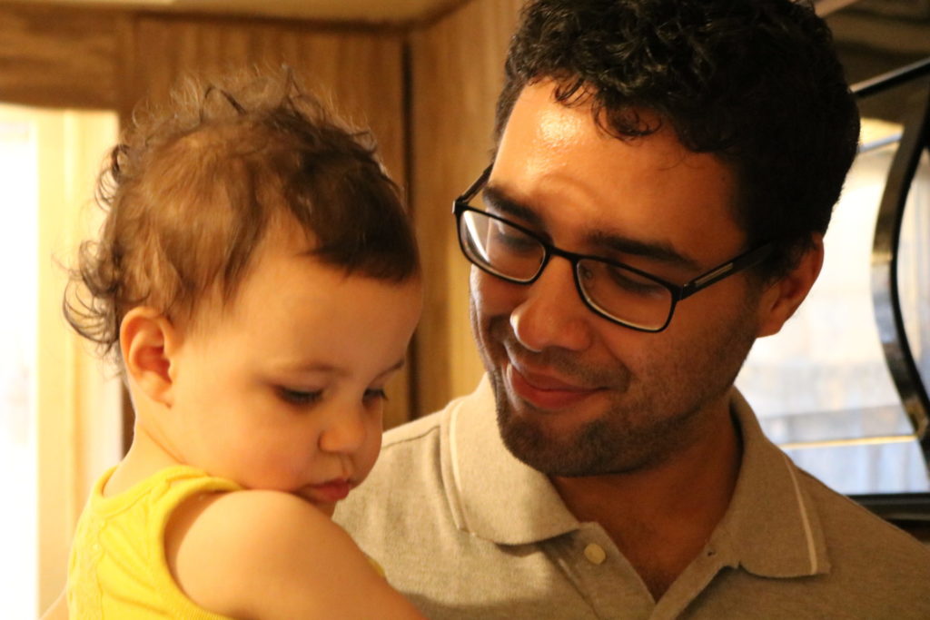 A man holding a toddler girl and smiling at her while the toddler looks down toward the ground