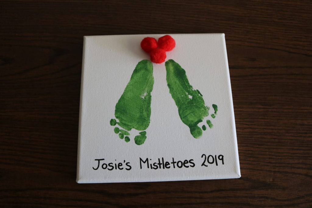 A white canvas with green baby footprints on it, red pom-poms, and the words "Josie's Mistletoes 2019"
