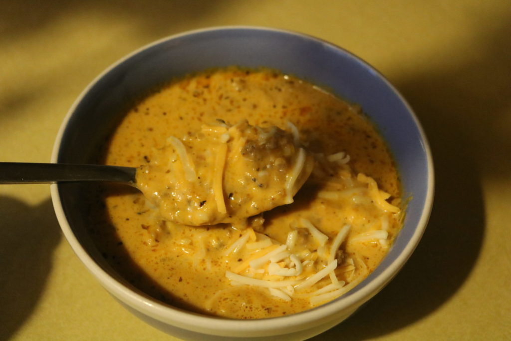 Soup topped with shredded cheese in a blue bowl, being scooped out by a spoon