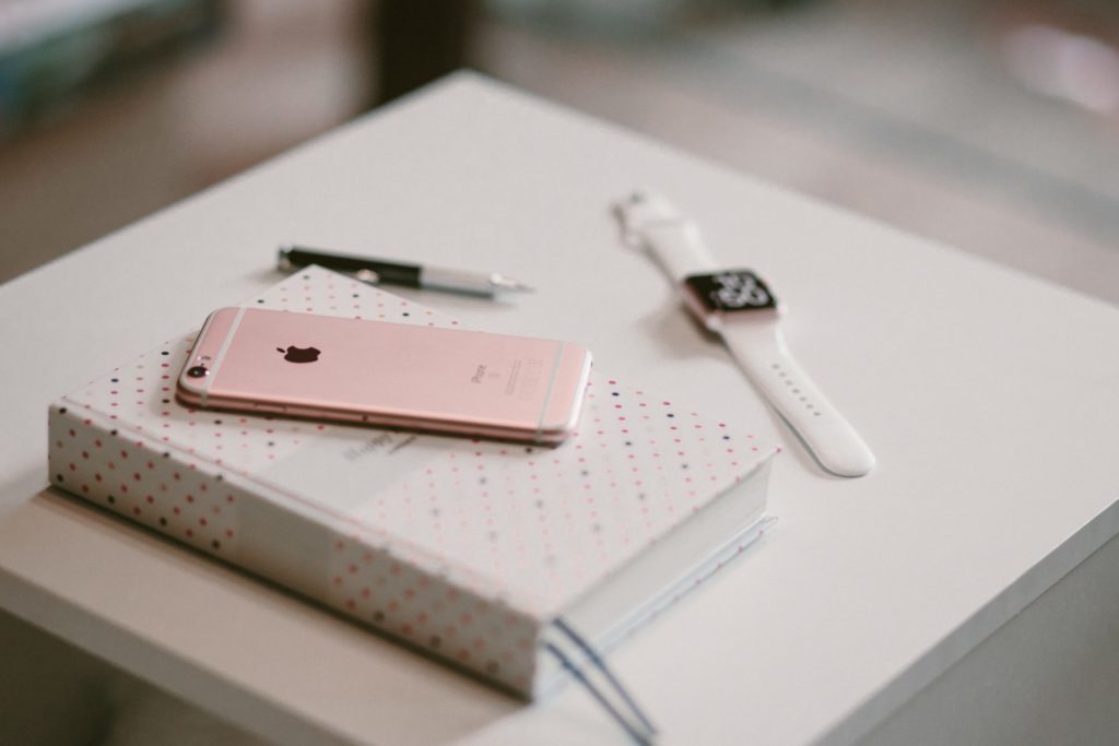 A pink iphone laying on top of a book beside an apple watch and a black pen