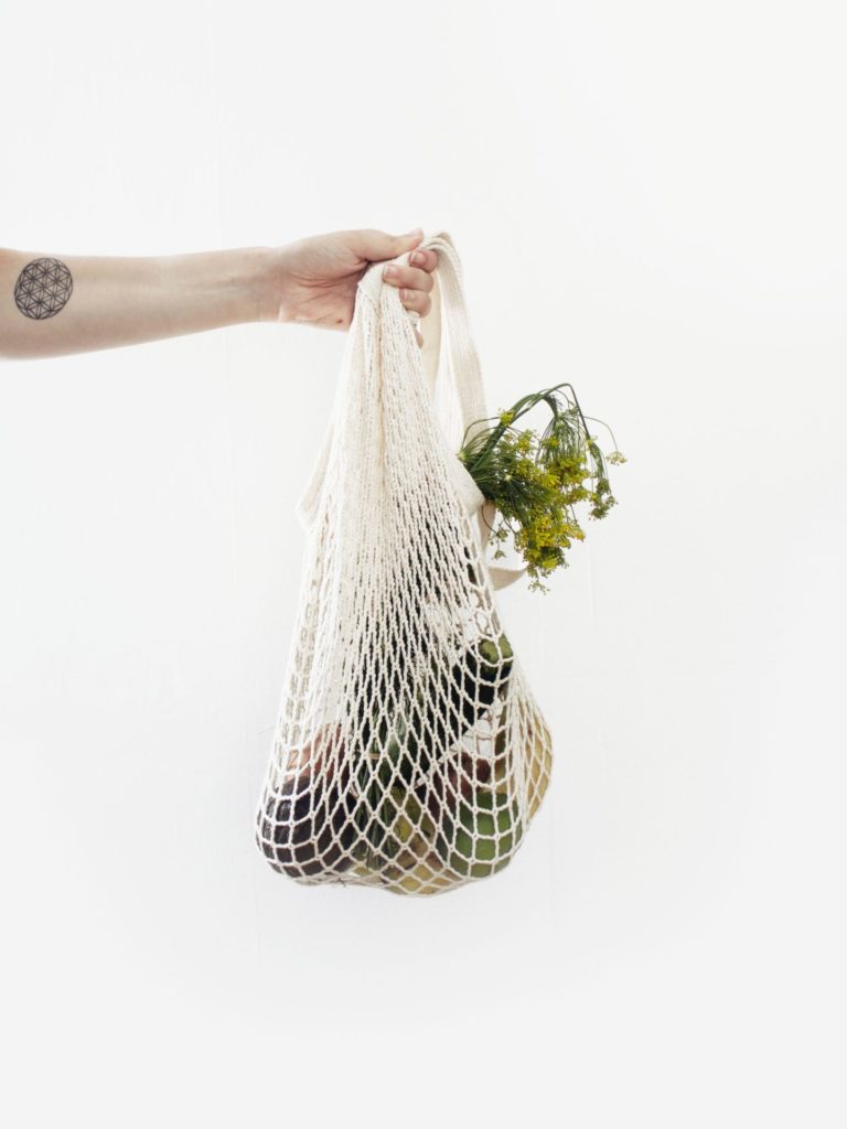 A tattooed arm holding out a reusable shopping bag containing produce